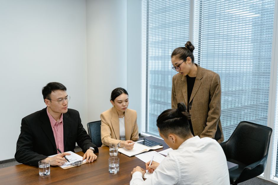 Four colleagues in a meeting room reviewing notes around a wooden table, with one standing to lead the discussion