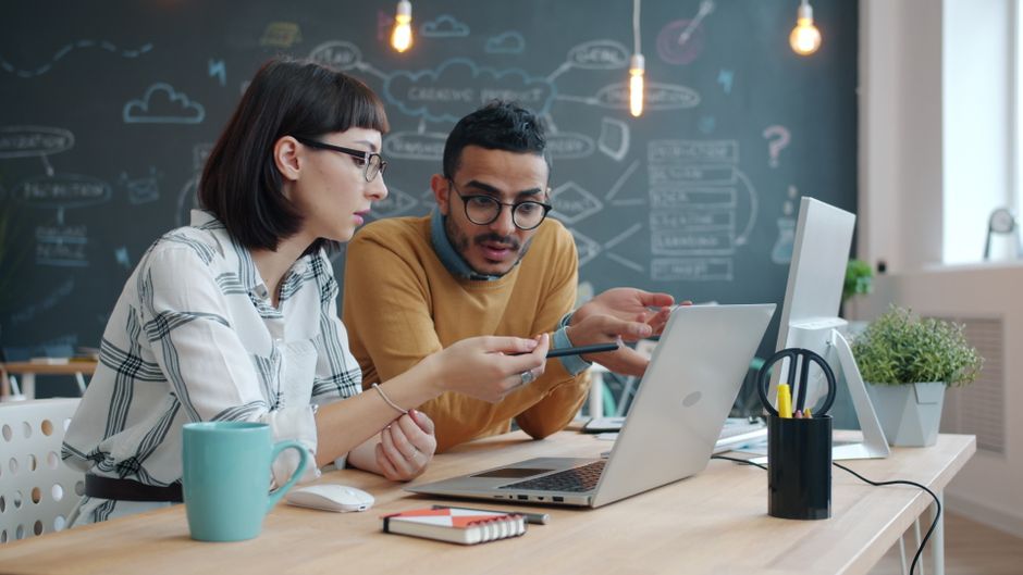 Two young founders working through plans on a laptop in front of a chalkboard covered in diagrams