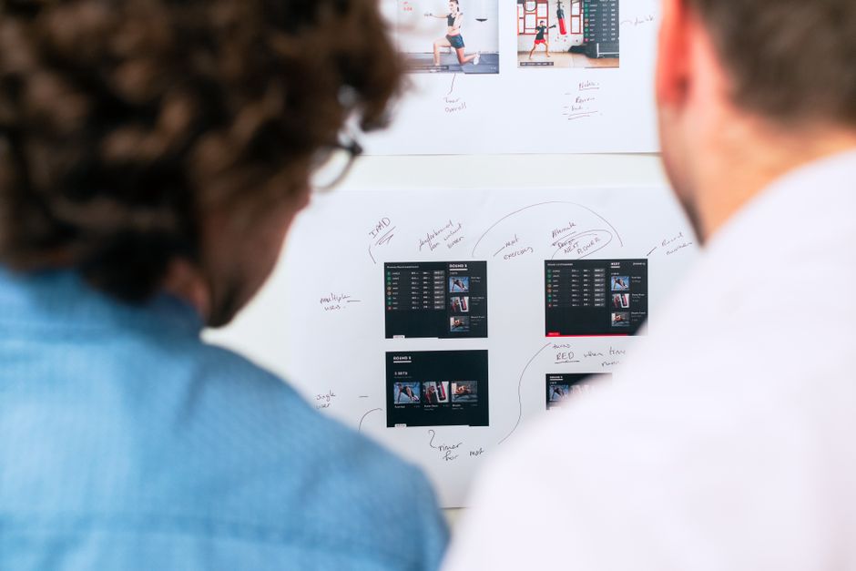 Two colleagues reviewing app screen designs and handwritten notes pinned to a wall while planning the next stage of a product