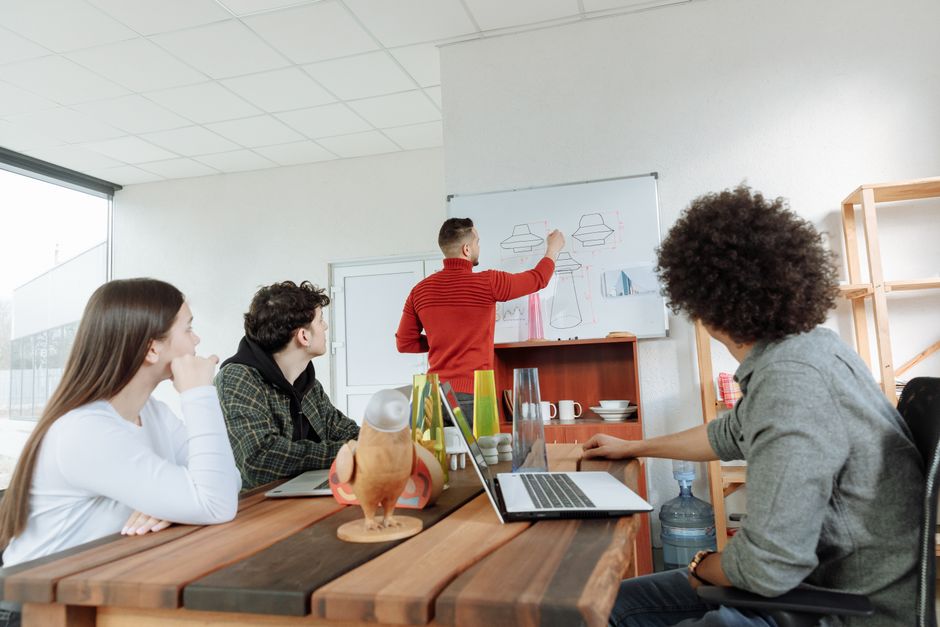 A mentor sketching ideas on a whiteboard for three young team members seated around a table with laptops